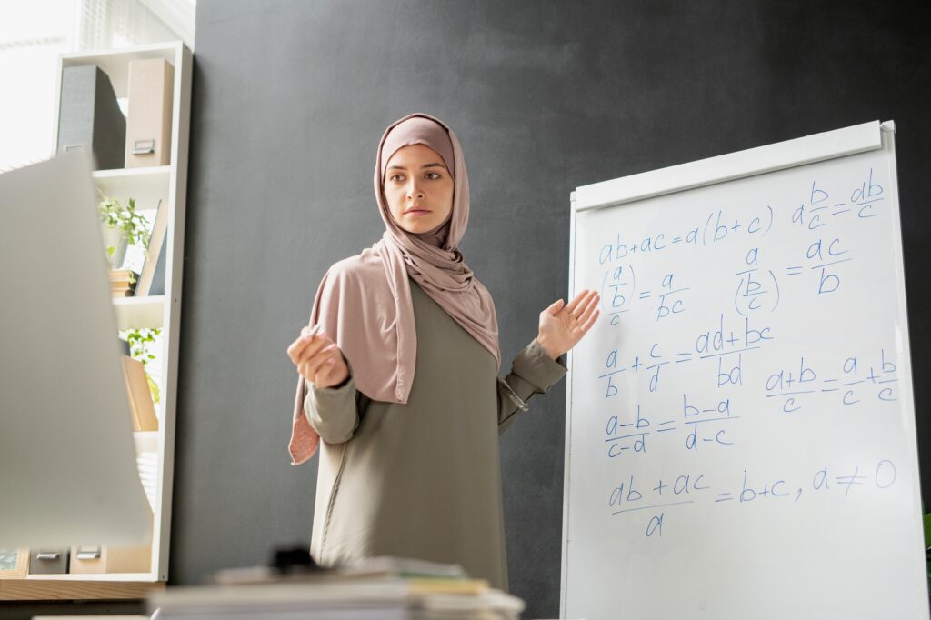 Young serious teacher of algebra standing by whiteboard in front of computer